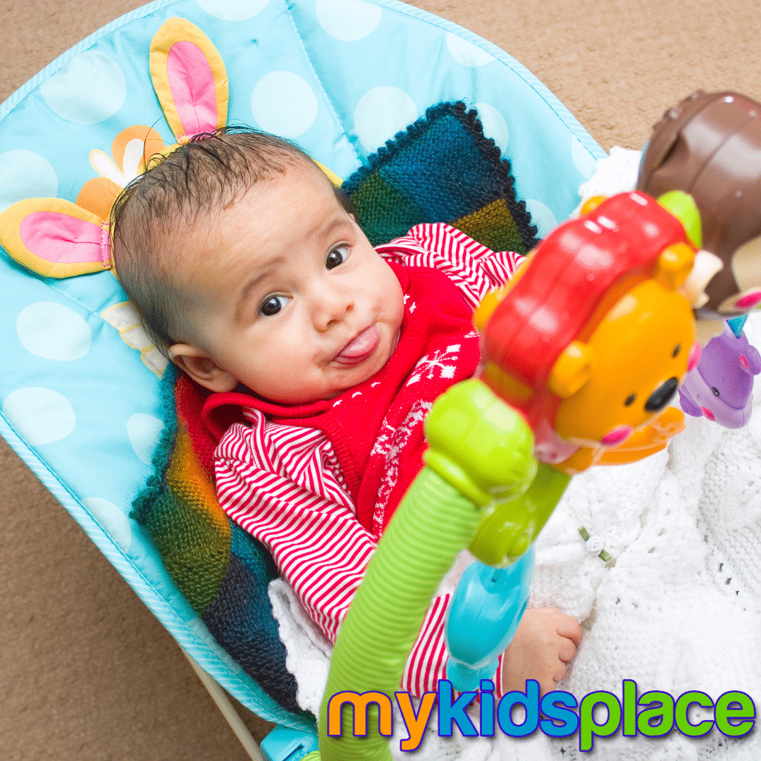 A baby in a red outfit lies down in a colorful rocker "container" while sticking tongue out at the camera.