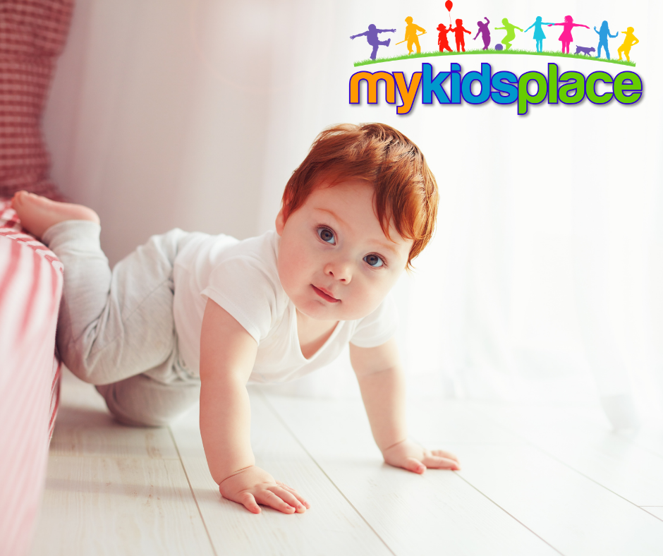 An infant with red hair wearing a white shirt and grey pants looks at the camera while creeps on hands and knees off of a low bed