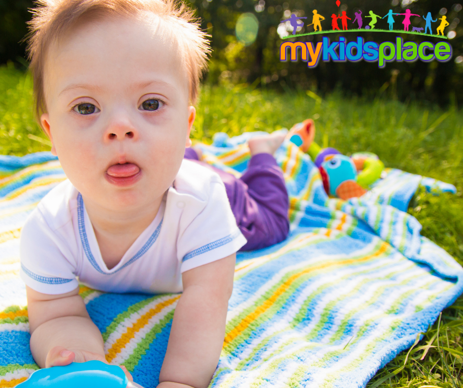 A baby with Down syndrome looks into the camera and practices tummy time on a multicolor striped blanket outside on the grass.