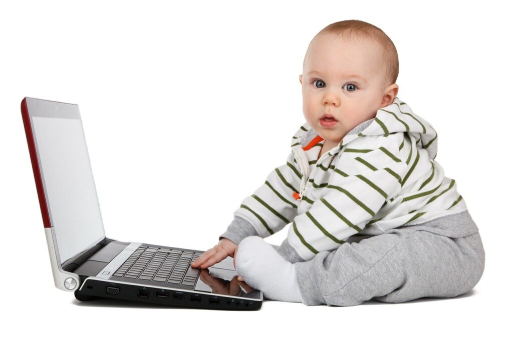 An infant in grey pants and a green-striped hoodie has their hand on a laptop as if ready for telehealth.