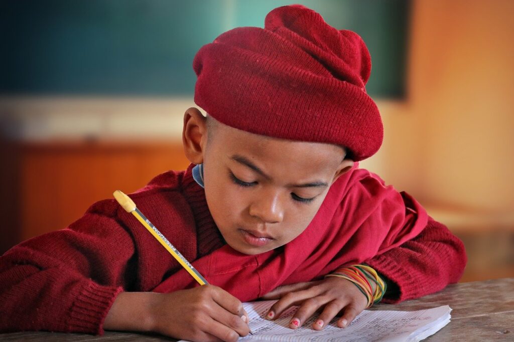 A school-aged child wearing a red hat and sweater is writing on lined paper with a pencil which is a useful fine motor skill.