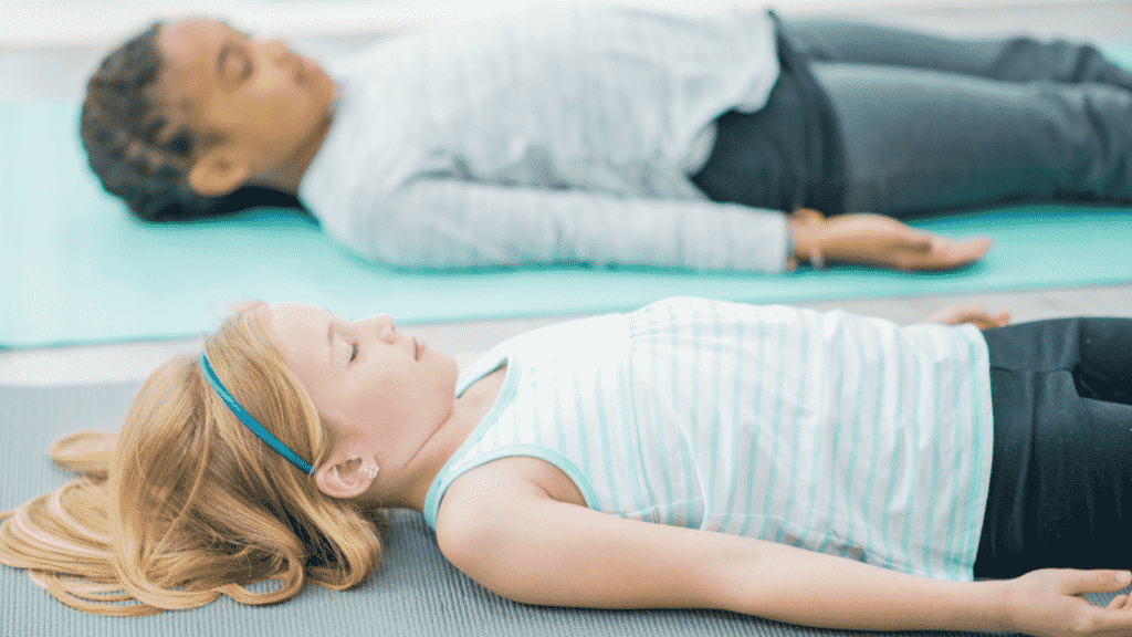 Two children lie on their backs, supine position, on yoga mats with eyes closed.