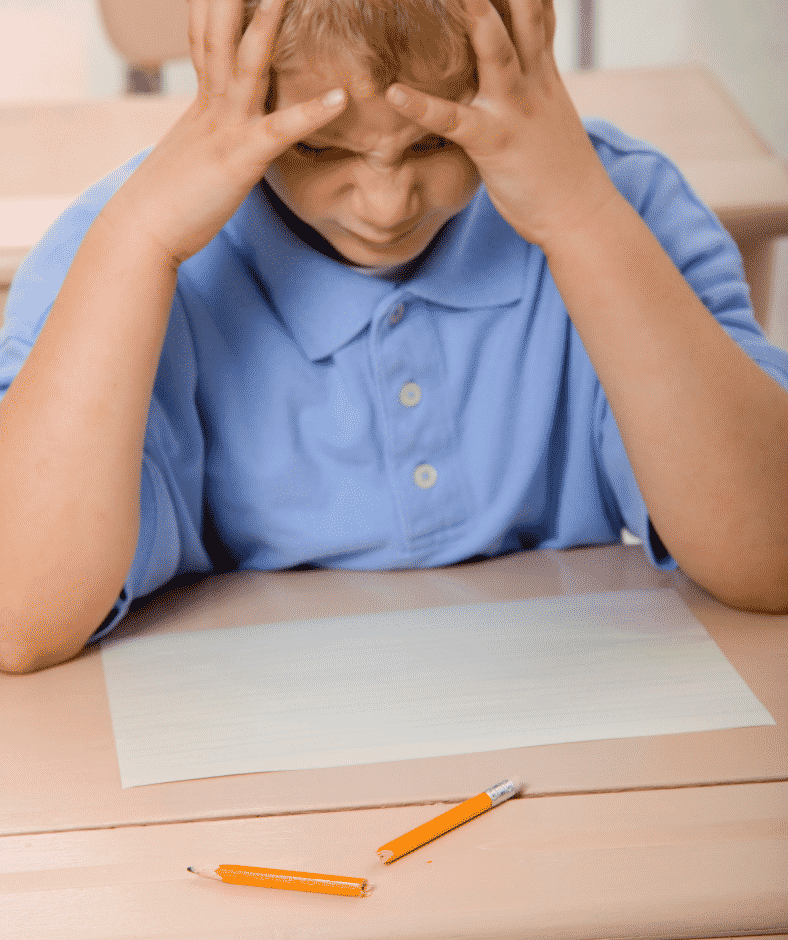 A frustrated child in a blue polo shirt has their hands on their face while they stare at a broken pencil which may have been the result of poor grading of force.