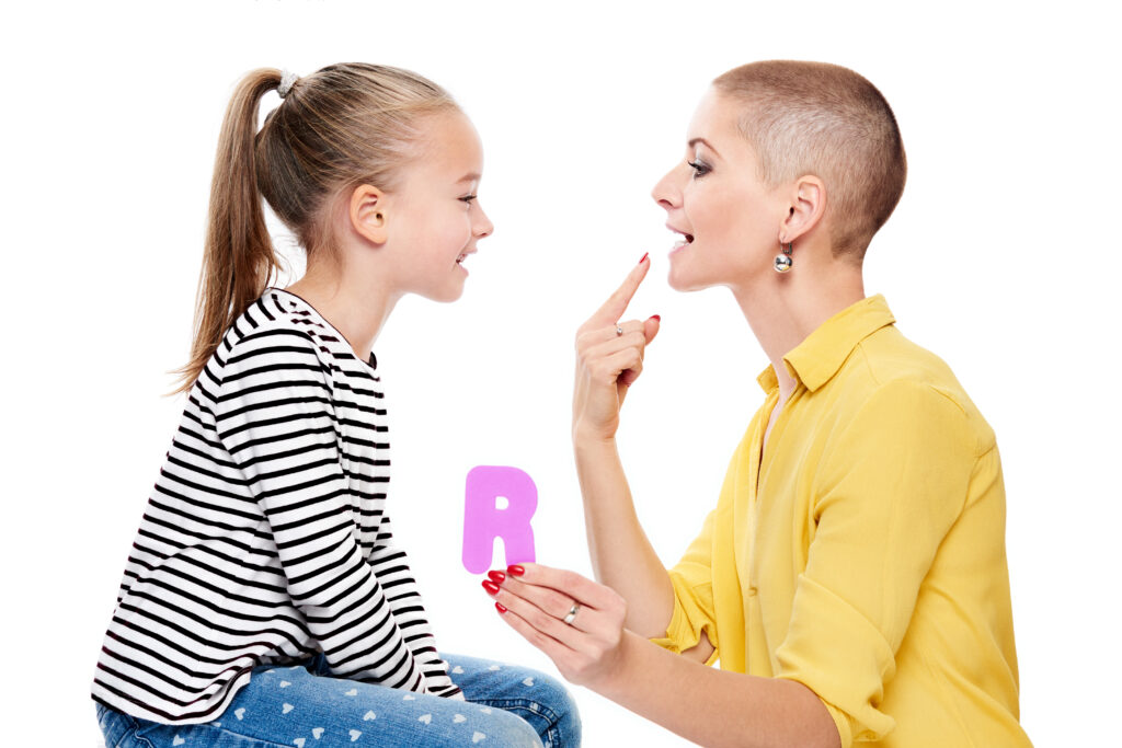 A speech therapist holds up a letter R while pointing to their mouth and forming the "r" sound. A child watches them closely and tries to replicate the sound.