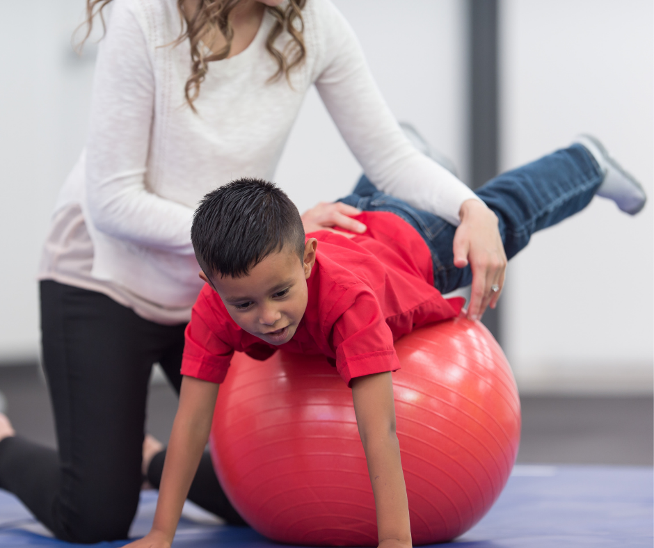 A child balances on an extra-large red ball face down using their hands on the floor as a physical therapist helps stabilize them at their hips.