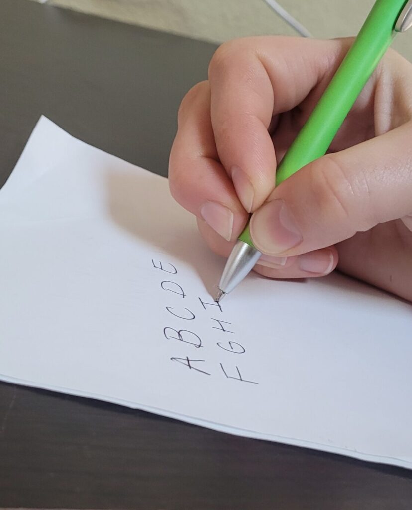A person with a quadrupod grasp on a green pen practices writing the alphabet in uppercase.