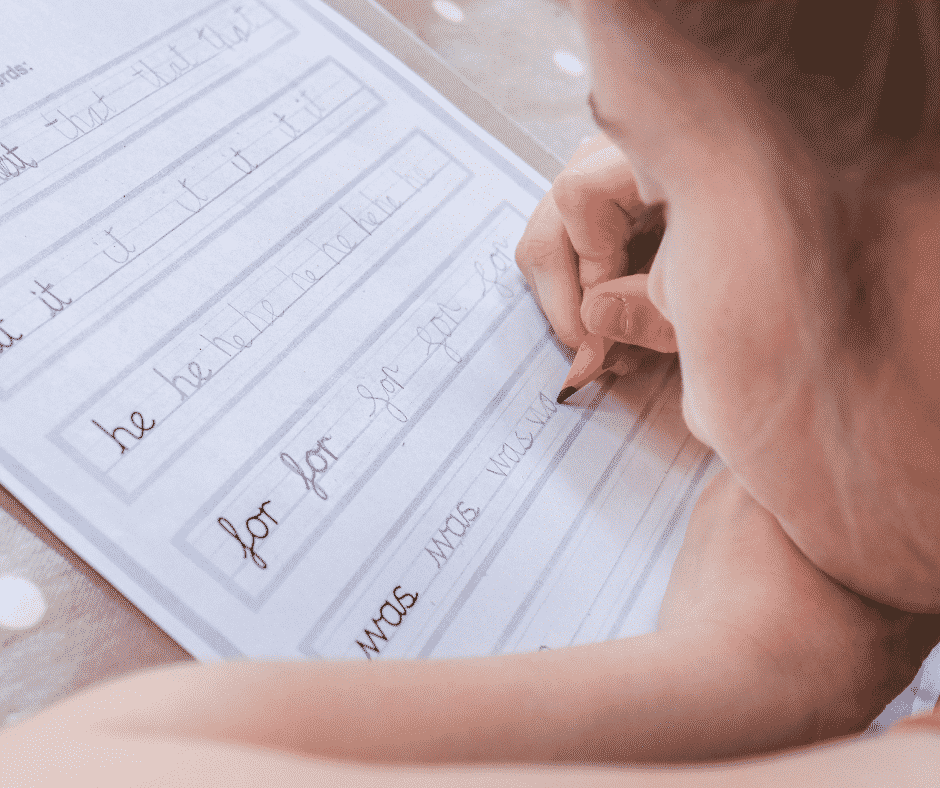A child practices writing in cursive and demonstrates a developing tripod grasp with their thumb and index finger on top of a pencil supported by their middle finger. The child rests their chin on their other hand.