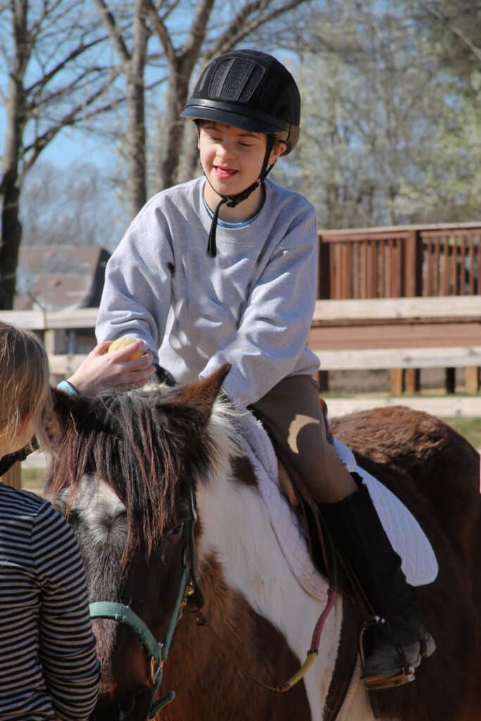 A child in a helmet is helped to ride a brown and white pony as a therapeutic intervention called hippotherapy.