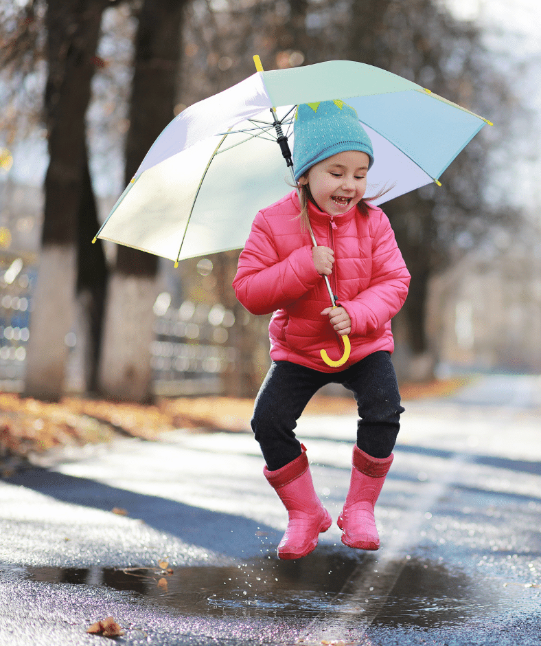A child with a blue umbrella, pink jacket, and pink rainboots is seen mid-air as they jump into a puddle.