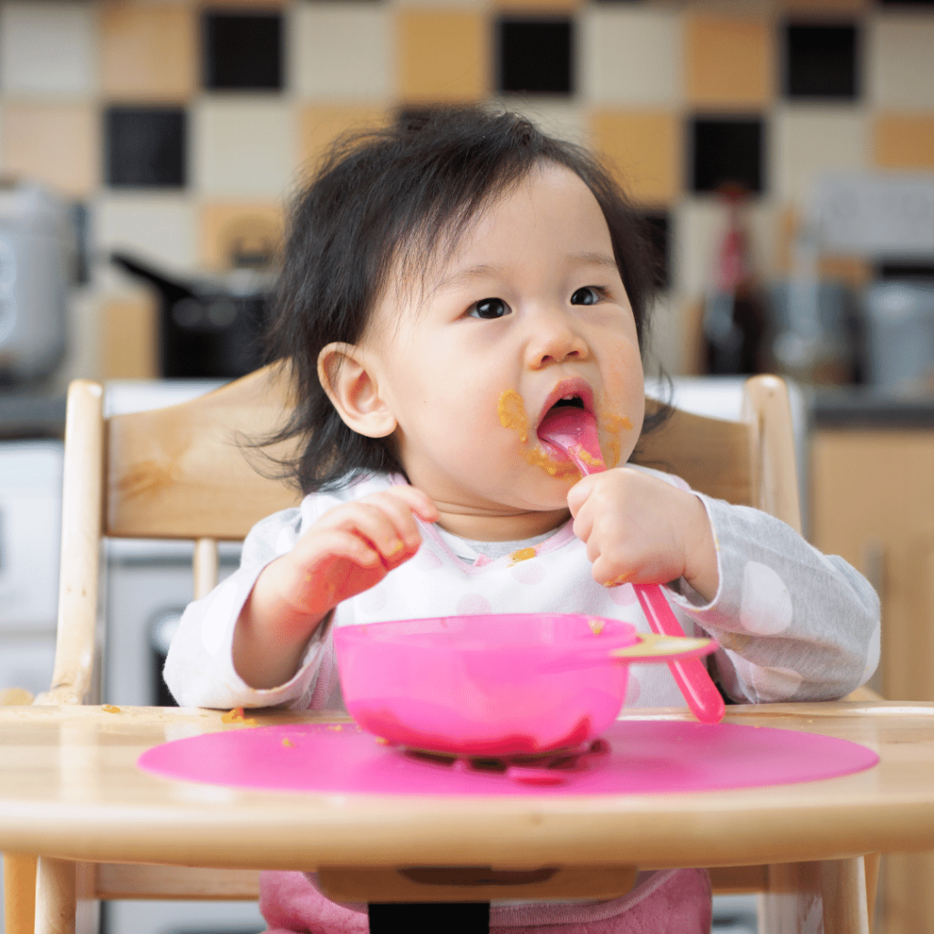 A baby with pureed food all over their mouth eats using a pink spoon while sitting in a high chair.
