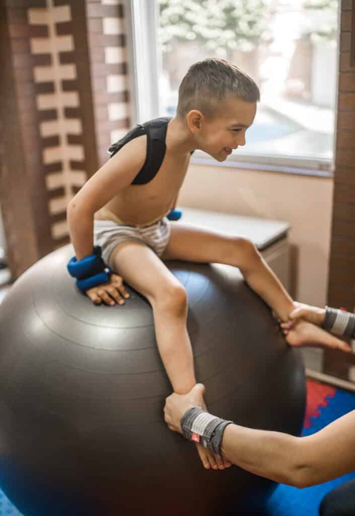 A smiling child wearing a postural support and wrist weights is stabilized on an extra-large therapy ball by an adult holding his legs as an exercise to improve postural stability.
