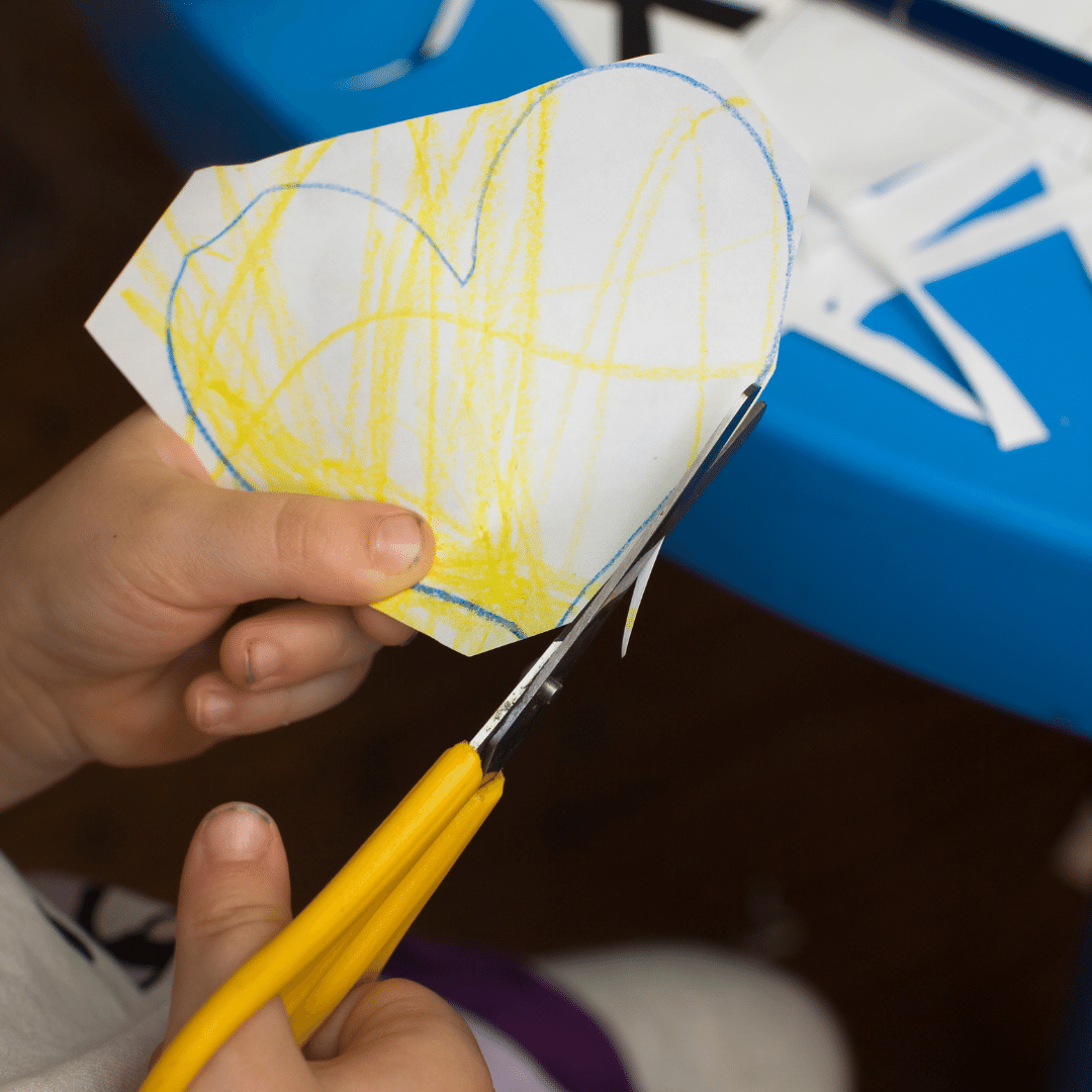Child's hands holding scissors attempt to cut out a heart outline that has been poorly colored in yellow. The yellow scribbles go outside the blue heart outline. The scissors missed the outline in several places.