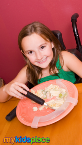 A smiling child in a wheelchair with long brown hair and wearing a green tank top uses an adaptive fork with a wide handle to eat pasta. An adaptive plate guard prevents food from being pushed off the plate and allows the child to pick up the food more easily.