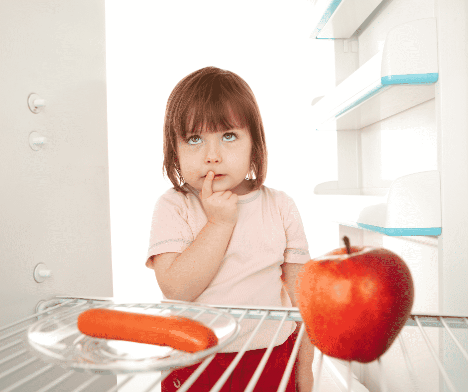 A child stands at an open refrigerator (facing the camera), holds their finger to their mouth and looks up as if deciding between a hotdog on a plate and a red apple.