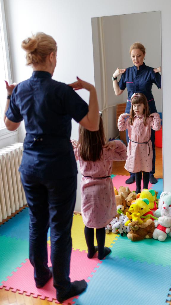 A child stands in front of a mirror with their hands on their shoulders while an adult stands behind them demonstrating the movement.